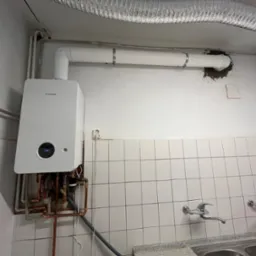 Nowoczesny, biały piec gazowy marki Junkers zamontowany na ścianie z białymi kafelkami, widoczne miedziane rury instalacji, biała rura odprowadzająca spaliny i fragment aluminiowego przewodu...