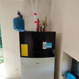 Nowa, częściowo owinięta folią, biało-czarna jednostka grzewcza Daikin z podłączonymi rurami i manometrem, zamontowana w surowym pomieszczeniu z oknem.