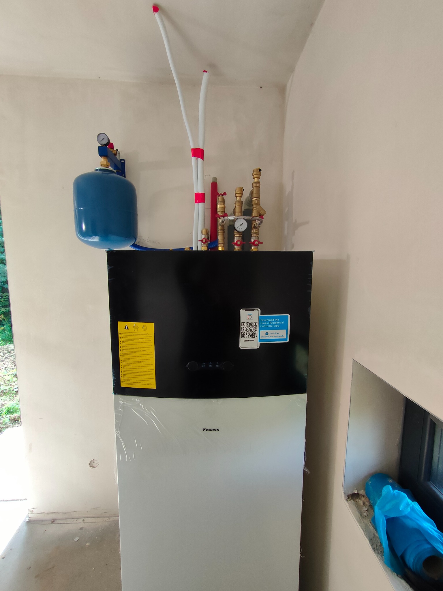 Nowa, częściowo owinięta folią, biało-czarna jednostka grzewcza Daikin z podłączonymi rurami i manometrem, zamontowana w surowym pomieszczeniu z oknem.