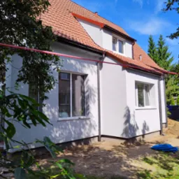 Jasnoszary dom jednorodzinny z czerwoną dachówką po remoncie elewacji, widoczny z boku, z elementami rusztowania i gruzem w tle.