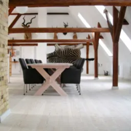 Jasna, bielona deska podłogowa lita w przestronnym, jasnym pokoju na poddaszu z drewnianymi belkami stropowymi, kolumną z cegły i dekoracjami w stylu myśliwskim.