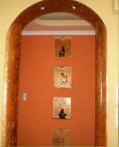 Pomarańczowa ściana z czterema obrazami w złotych ramach, widoczna przez ozdobny łuk z efektem drewna.