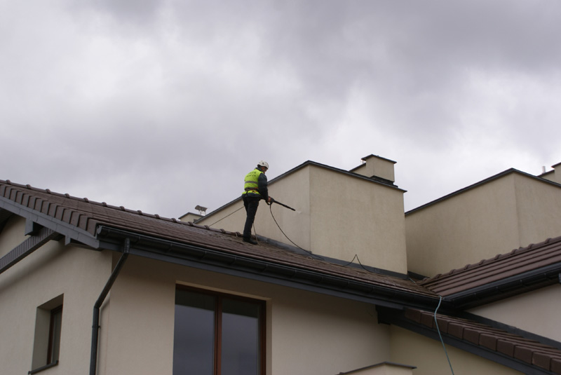 Pracownik w kasku i kamizelce odblaskowej myje dach domu za pomocą myjki ciśnieniowej, widoczne brązowe dachówki i beżowa elewacja budynku na tle pochmurnego nieba.
