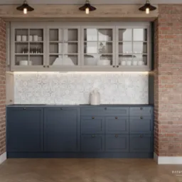 Granatowa zabudowa kuchenna z frezowanymi frontami i mosiężnymi gałkami, uzupełniona o szklane szafki wiszące i ceglaną ścianę, oświetlona trzema lampami w stylu industrialnym.