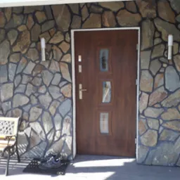 Wejście do domu z brązowymi drzwiami z szybkami, wykończone kamienną elewacją w odcieniach szarości i brązu, oświetlone dwoma lampami na ścianie, z ławką i butami na wycieraczce.