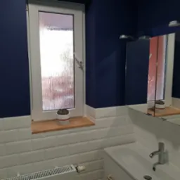 Niebieska ściana w łazience z białymi, prostokątnymi płytkami, biała umywalka z szafką, okno z ozdobną szybą i drewnianym parapetem z doniczką, odbicie w lustrze