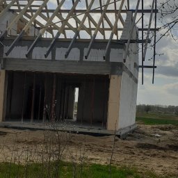 Buzdys Usługi Budowlane - Surowy stan domu z białych bloczków z drewnianą konstrukcją dachu, widok z zewnątrz na tle zachmurzonego nieba i polnego krajobrazu.