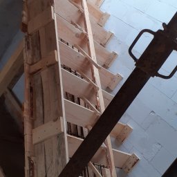 Buzdys Usługi Budowlane - Szczegółowe ujęcie drewnianego szalunku schodów z widocznym zbrojeniem, wspartego metalowymi stemplami, na tle ściany z bloczków betonowych.