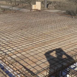 Zbrojenie płyty fundamentowej z widocznym cieniem osoby fotografującej, drewniane szalunki i izolacja termiczna w tle.