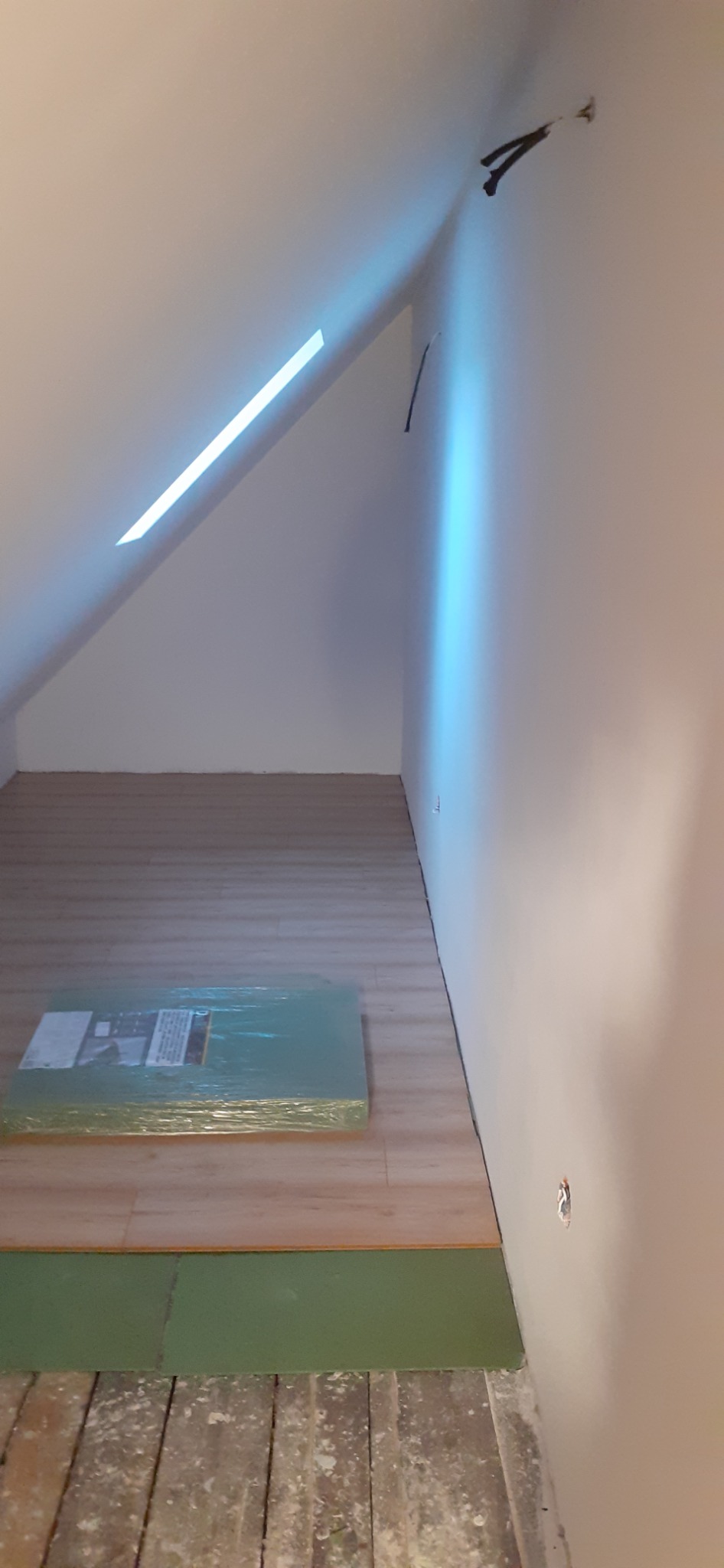 Poddasze w trakcie remontu: położona podłoga z paneli, paczka paneli w folii leżąca na podłodze, widoczne stare deski pod spodem, skośny sufit z oknem dachowym.