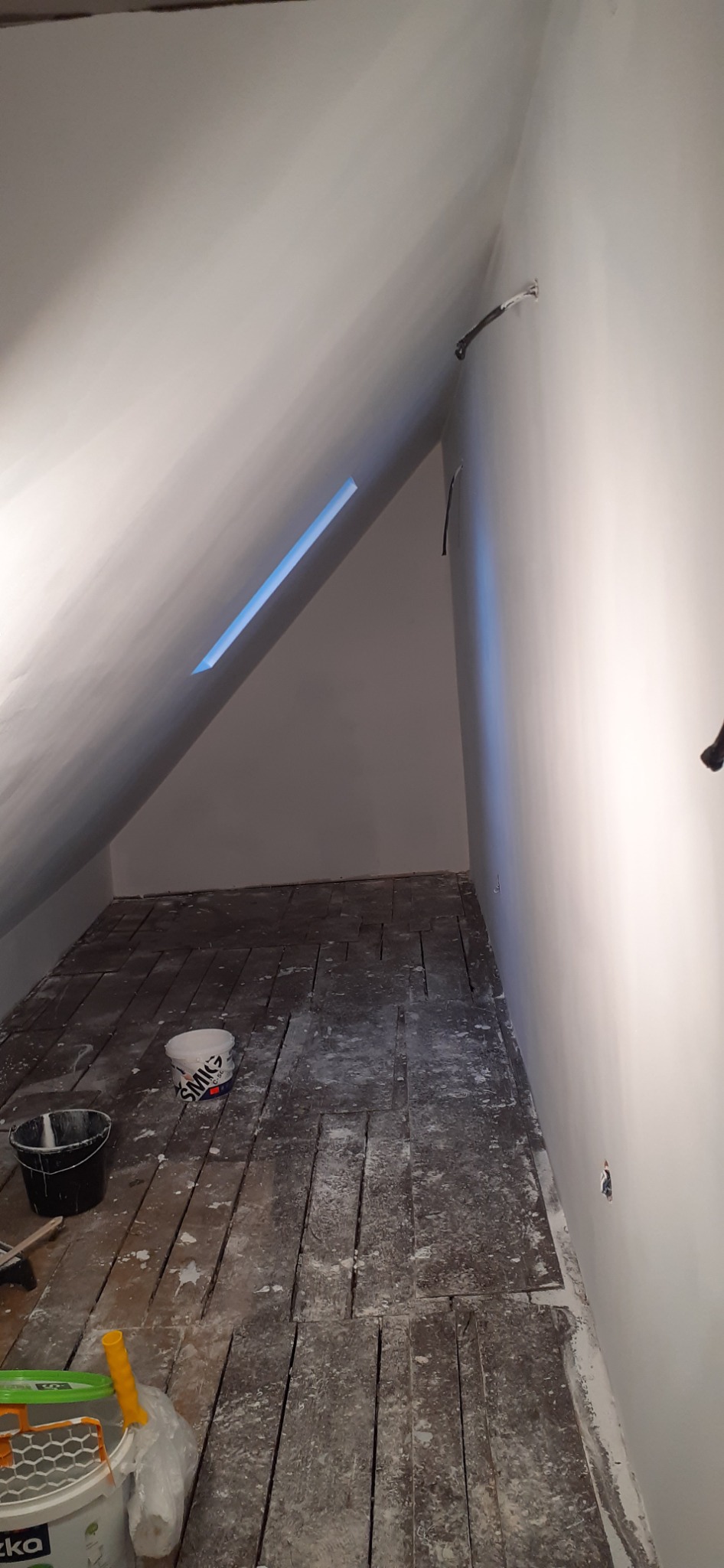 Poddasze po malowaniu ścian na biało, widoczne drewniane deski podłogowe pokryte farbą, wiadro z farbą i wałek malarski w rogu, małe okno dachowe w skosie.