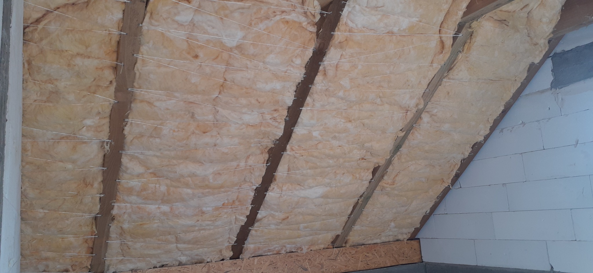 Ocieplenie poddasza wełną mineralną, zabezpieczone sznurkiem, widoczne drewniane krokwie i fragment ściany z białych bloczków.