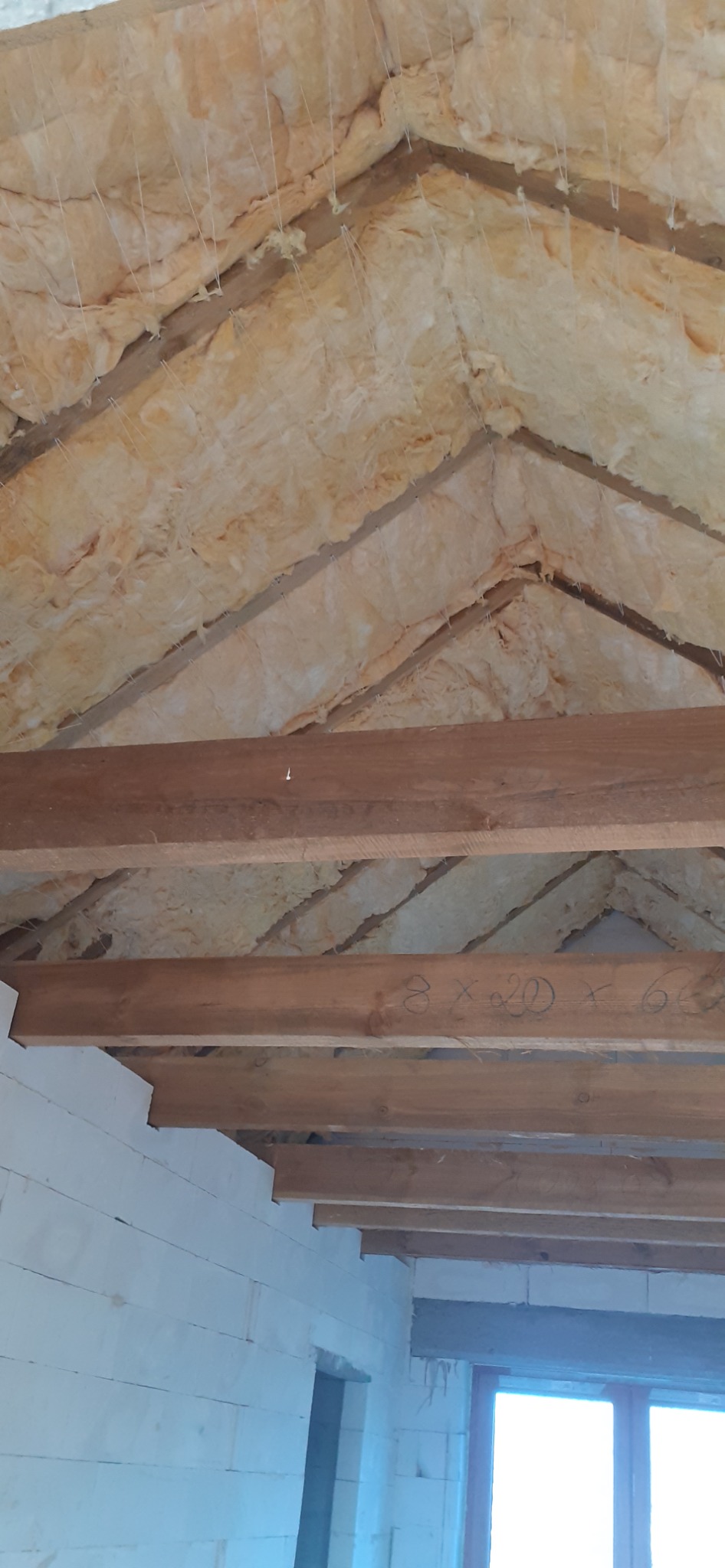 Izolacja poddasza wełną mineralną, widoczne drewniane krokwie i sznurki mocujące, ściany z białego betonu komórkowego.