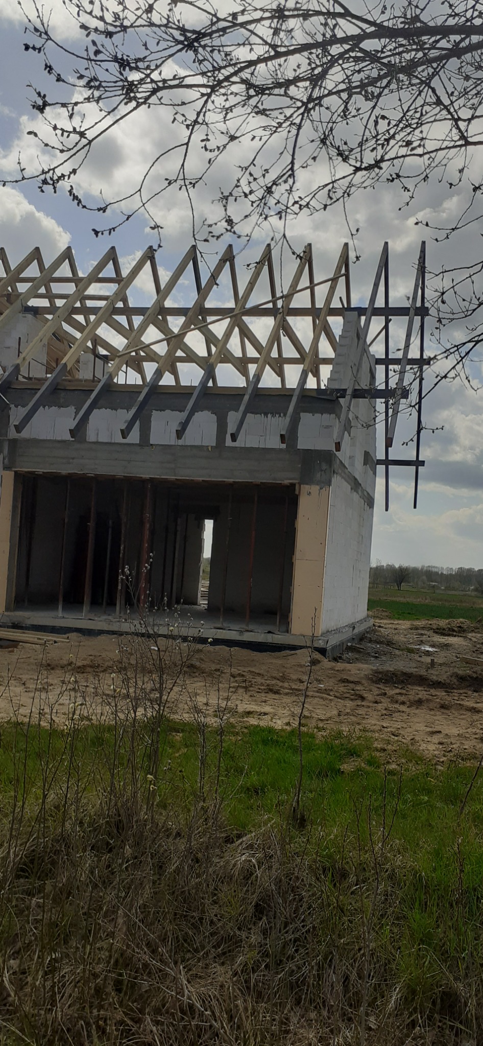 Surowy stan domu z białych bloczków z drewnianą konstrukcją dachu, widok z zewnątrz na tle zachmurzonego nieba i polnego krajobrazu.