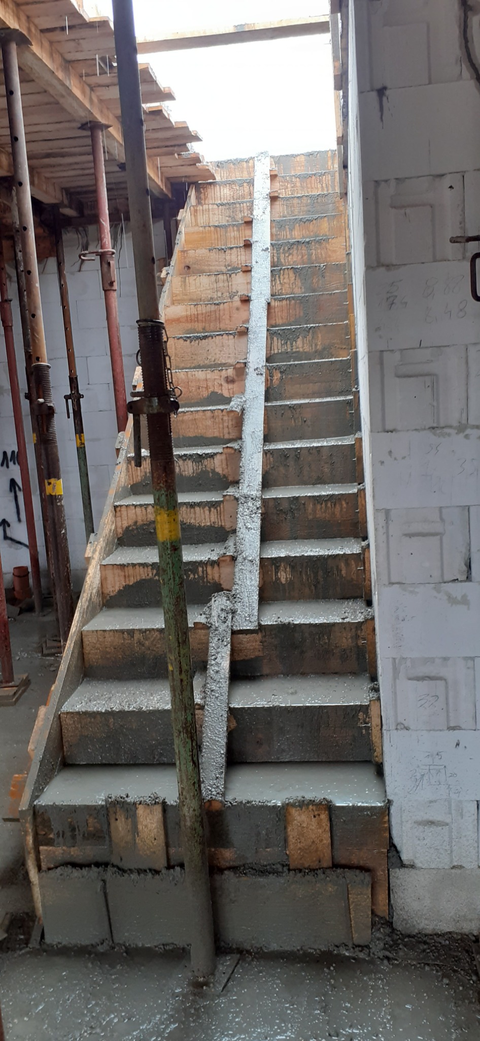 Betonowe schody w trakcie budowy, widoczne szalunki i podpory, ściana z bloczków betonowych z naniesionymi notatkami.