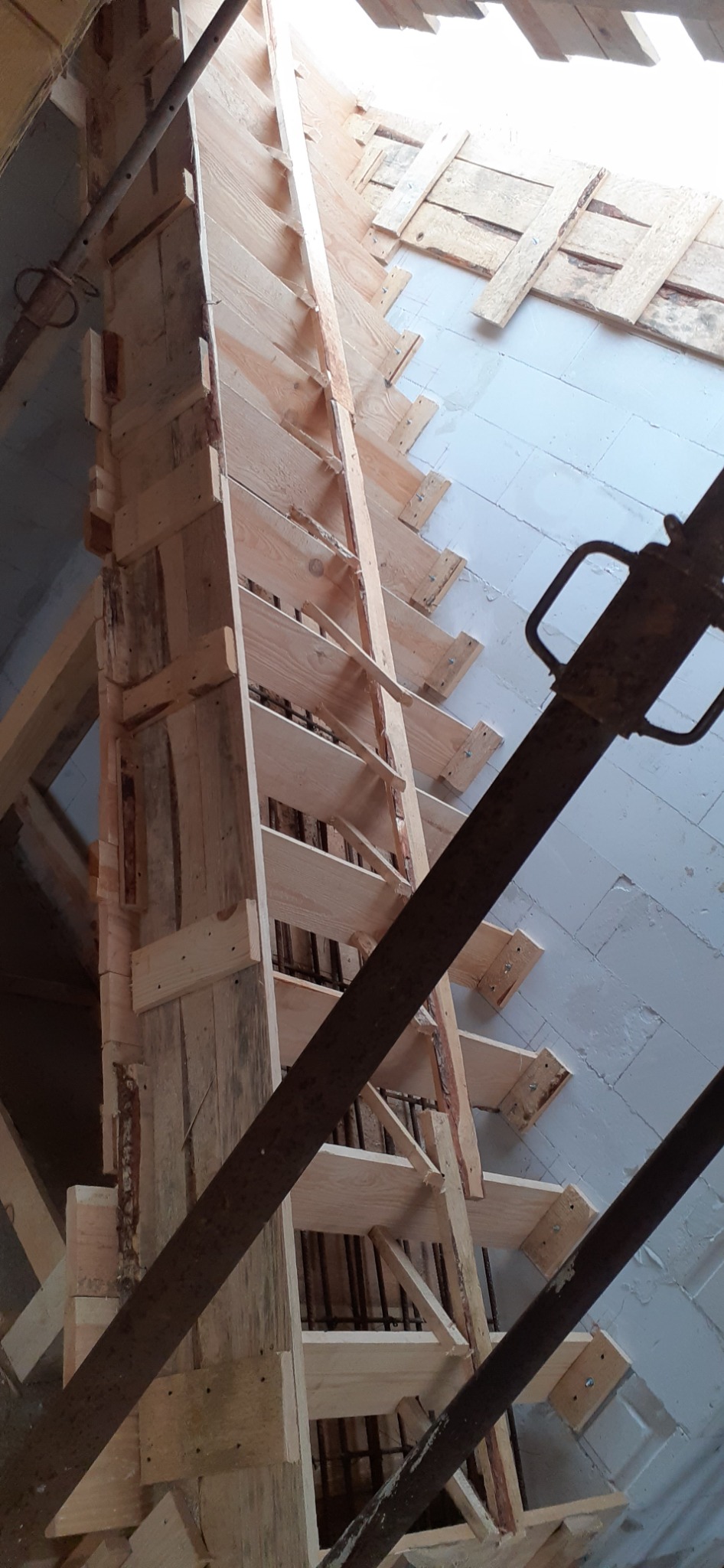 Szczegółowe ujęcie drewnianego szalunku schodów z widocznym zbrojeniem, wspartego metalowymi stemplami, na tle ściany z bloczków betonowych.
