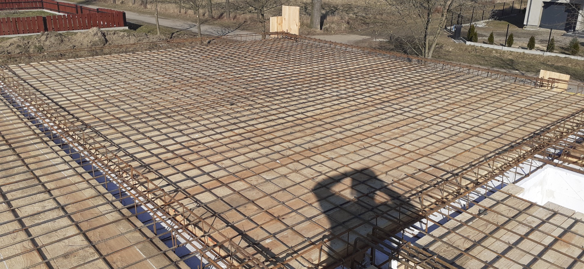 Zbrojenie płyty fundamentowej z widocznym cieniem osoby fotografującej, drewniane szalunki i izolacja termiczna w tle.