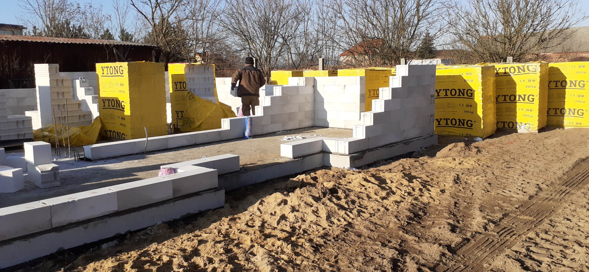 Budowa domu z bloczków Ytong, widoczne zapakowane palety bloczków, częściowo wzniesione ściany fundamentowe i pracownik w środku konstrukcji.
