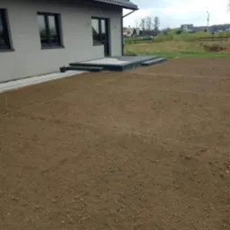 Przygotowany teren pod przyszły trawnik z wytyczonymi liniami, widoczny fragment domu z tarasem i schodami.