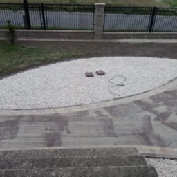 Ogród z wysypanym białym żwirem, obok ułożona kostka brukowa w odcieniach brązu i szarości, całość otoczona niskim murkiem i czarnym metalowym płotem.