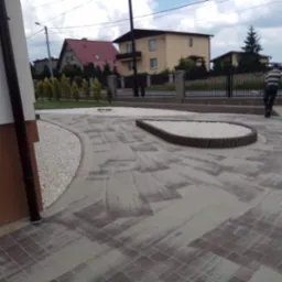 Ułożona kostka brukowa z dekoracyjnymi obrzeżami z palisady i białego grysu na podjeździe przed domem jednorodzinnym, w tle osoba przechodząca przez bramę.