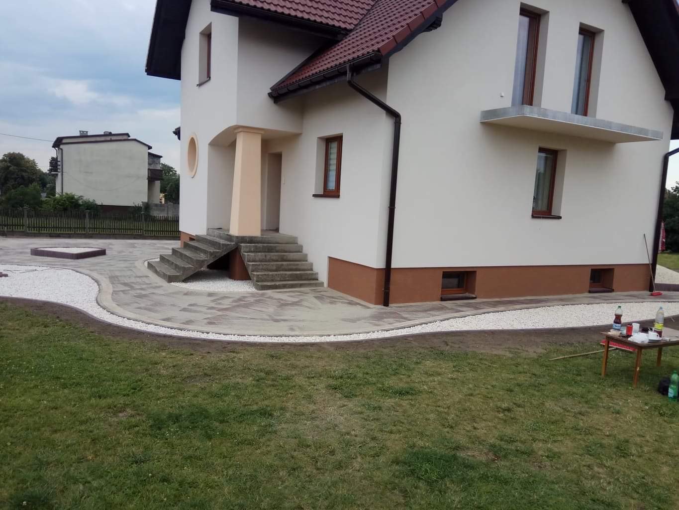 Ułożona szara kostka brukowa przed domem z białym kamieniem ozdobnym wzdłuż krawędzi, schody wejściowe z kostki, trawnik w pierwszym planie.