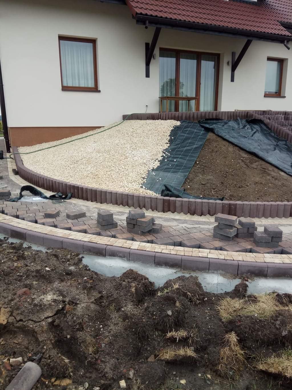 Kaskadowy ogród przed domem w trakcie realizacji: ułożona obrzeża z kostki, wysypany biały kamień, rozłożona czarna agrowłóknina, przygotowane kostki do dalszego układania.