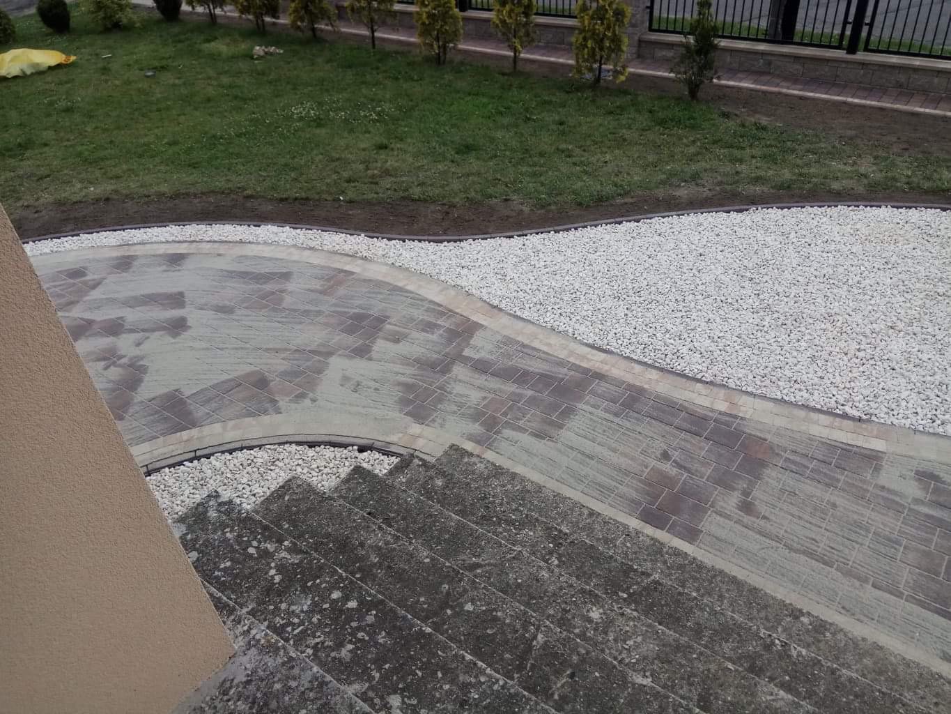 Ścieżka z kostki brukowej w odcieniach szarości i brązu, obrzeża z jasnego żwiru, widok z góry na schody i fragment budynku.