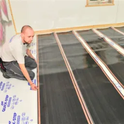 Montaż folii grzewczej podłogowej Quick-Step przez instalatora; widoczne paski grzewcze z miedzianymi przewodami na podkładzie izolacyjnym, narzędzia instalacyjne w kaburze przy pasie montera.