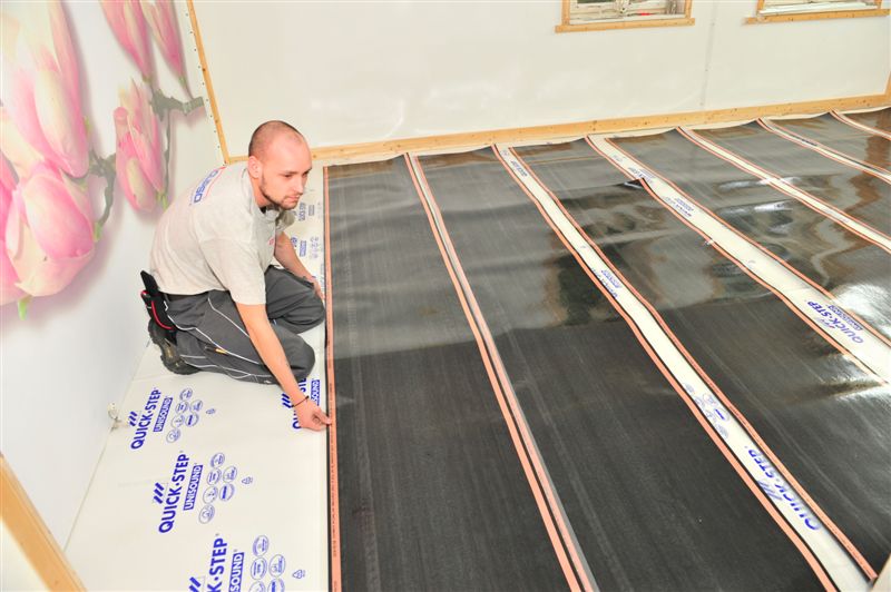 Montaż folii grzewczej podłogowej Quick-Step przez instalatora; widoczne paski grzewcze z miedzianymi przewodami na podkładzie izolacyjnym, narzędzia instalacyjne w kaburze przy pasie montera.