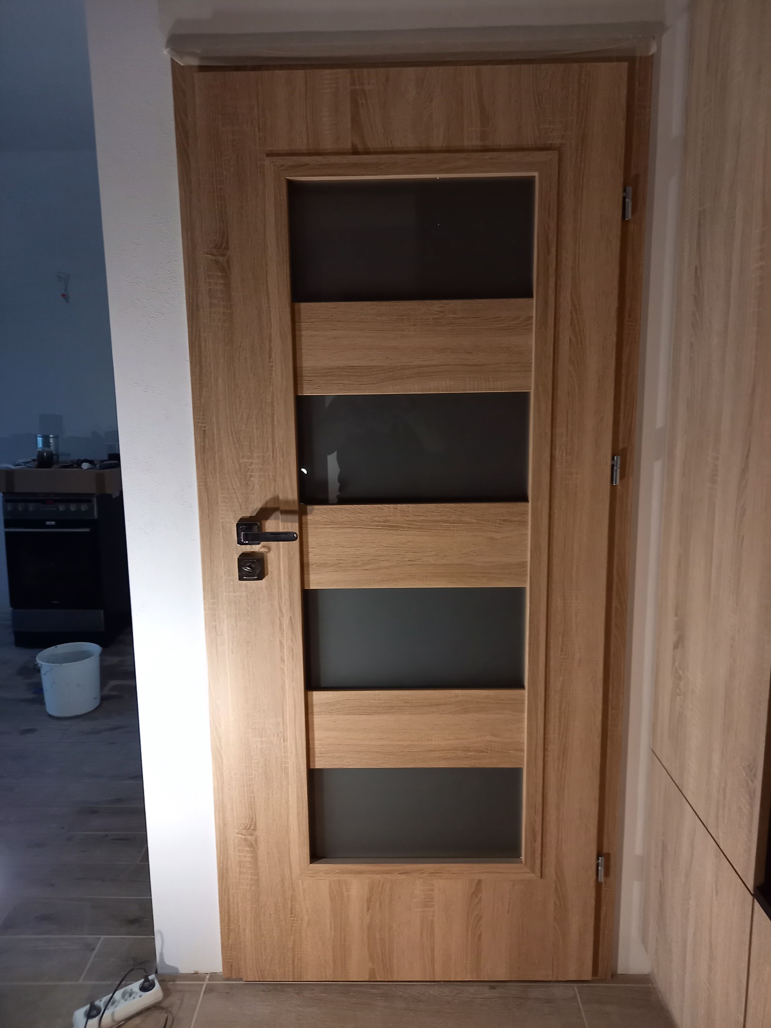 Zamknięte drzwi wewnętrzne z drewnianą ramą i czterema poziomymi szybami z matowego szkła, widoczne w trakcie montażu w pomieszczeniu z płytkami podłogowymi i sprzętem kuchennym w tle.