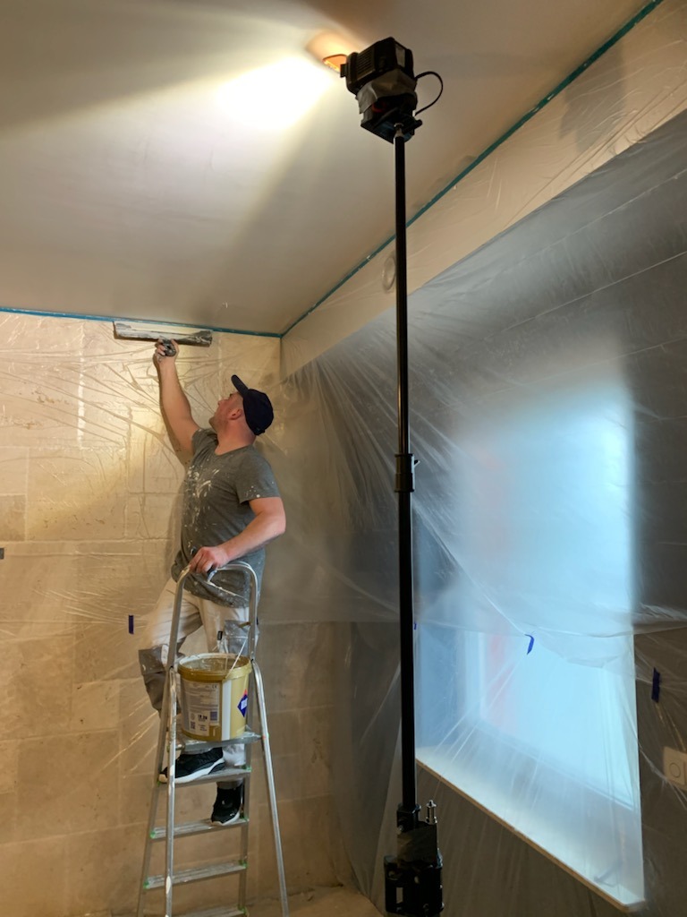 Mężczyzna na drabinie szpachluje sufit w pomieszczeniu zabezpieczonym folią malarską, oświetlonym lampą na wysięgniku.