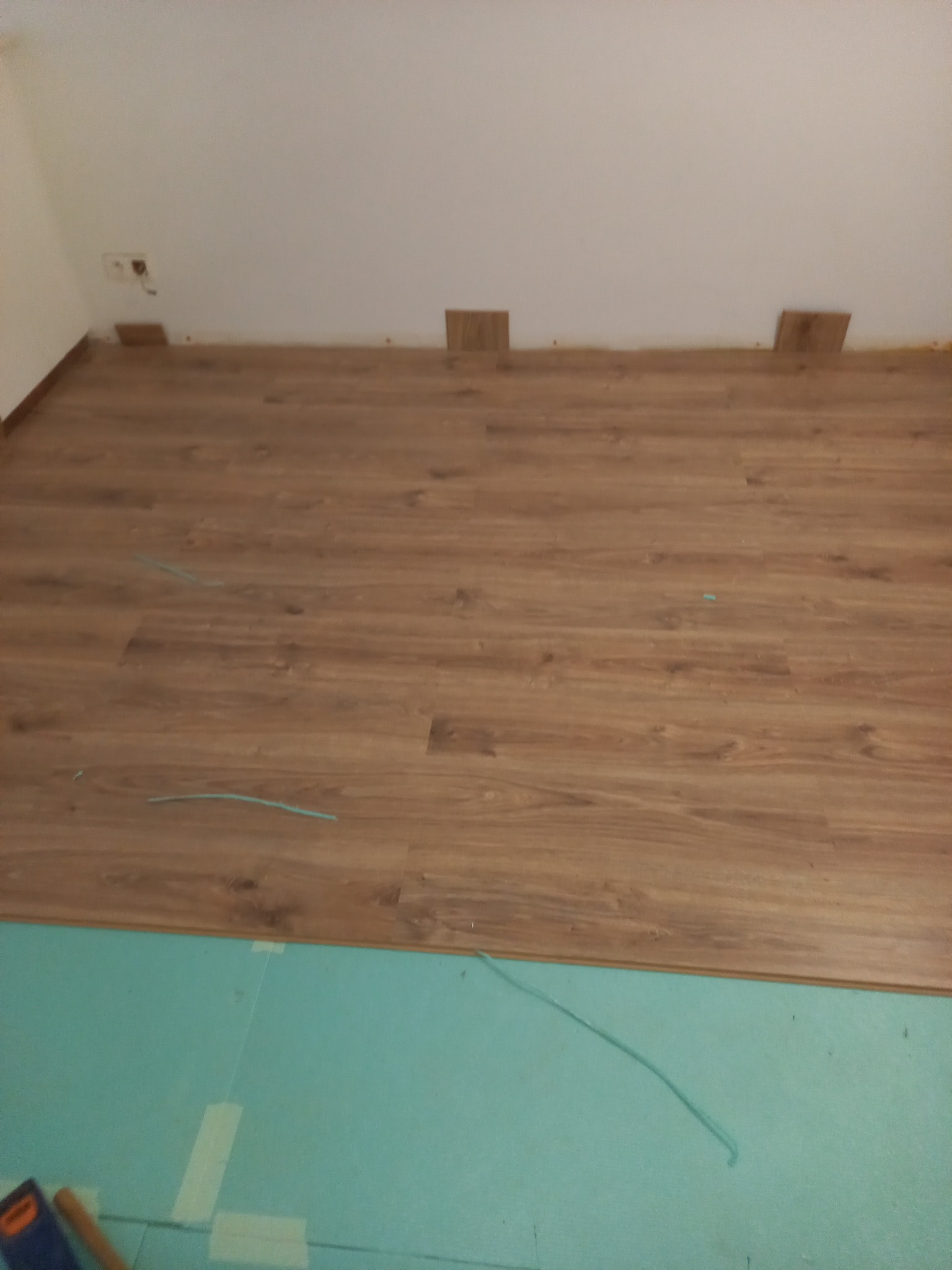 Proces układania paneli podłogowych w pomieszczeniu, widoczna podkładka izolacyjna i fragment narzędzi.