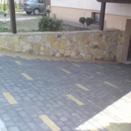 Ułożona kostka brukowa w odcieniach szarości i beżu, tworząca wzór na podjeździe. W tle kamienny murek z elementami piaskowca i fragment budynku z oknem oraz garażem.