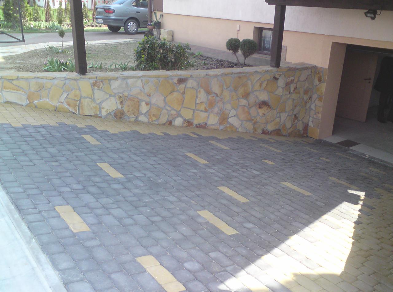 Ułożona kostka brukowa w odcieniach szarości i beżu, tworząca wzór na podjeździe. W tle kamienny murek z elementami piaskowca i fragment budynku z oknem oraz garażem.