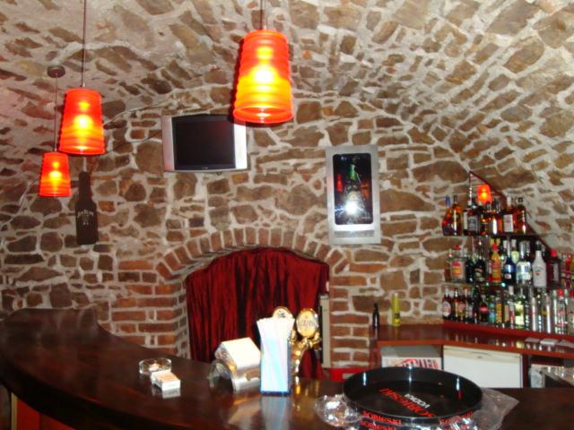 Kamienna ściana i sklepienie w barze z oświetleniem, telewizorem i półkami z alkoholami. Czerwone zasłony w tle. Blat baru z akcesoriami.