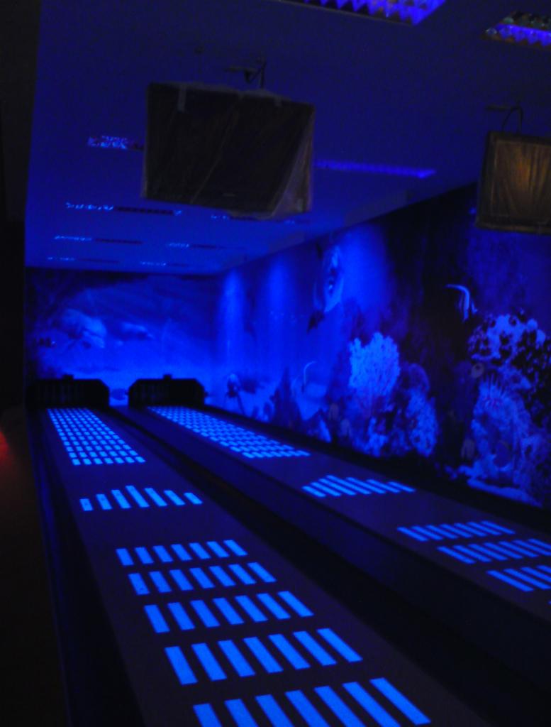 Oświetlone toru do gry w kręgle z motywem oceanu na ścianie, podświetlone niebieskim światłem. Widoczne dwa tory i elementy dekoracyjne.