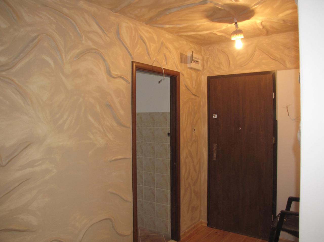 Beżowe ściany z dekoracyjną fakturą imitującą fale, drewniane drzwi wejściowe i otwarte drzwi do łazienki z beżowymi płytkami. Widok na wnętrze mieszkania.