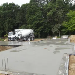 Świeżo wylany fundament pod budowę domu, z widocznym betonowozem w tle i wystającymi prętami zbrojeniowymi.