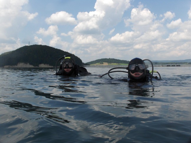 Dwóch nurków w pełnym sprzęcie na tle spokojnej wody, zalesionego wzgórza i błękitnego nieba z chmurami.