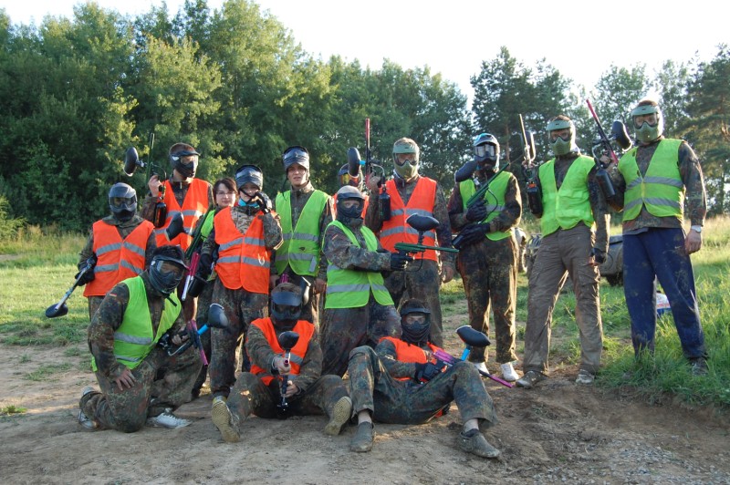 Grupa osób w maskach ochronnych i kamizelkach, trzymających markery paintballowe, pozuje na tle lasu.