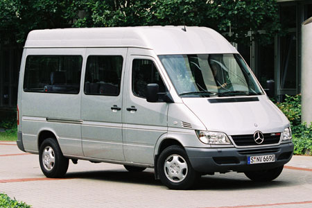 Srebrny bus marki Mercedes-Benz Sprinter z wysokim dachem, zaparkowany na brukowanym placu przed budynkiem z ciemnozielonymi krzewami w tle.