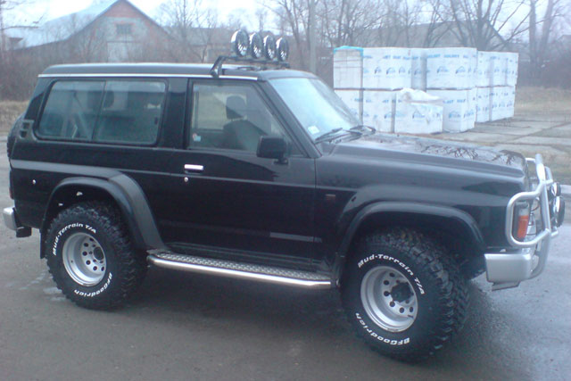 Czarny, dwudrzwiowy terenowy samochód z dodatkowym oświetleniem na dachu i oponami terenowymi, zaparkowany na zewnątrz, w tle budynek gospodarczy i sterta materiałów budowlanych.