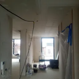 Wnętrze w trakcie prac: ściany i sufit po nałożeniu gładzi, widoczne worki z gładzią, kable elektryczne, okno z widokiem na budynek, podłoga zakurzona, zabezpieczona folią część ściany.