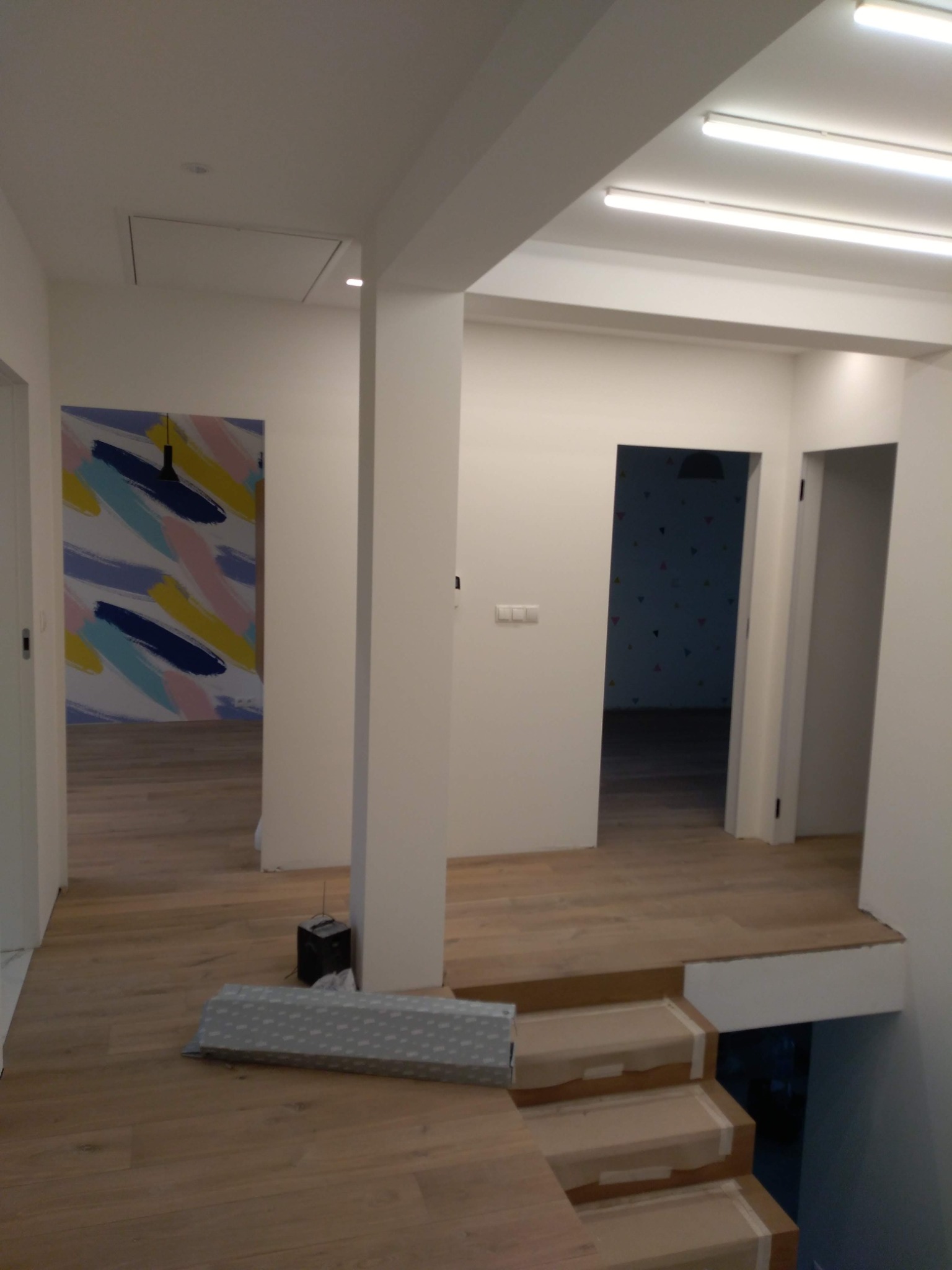 Wnętrze w trakcie prac wykończeniowych: drewnopodobne panele ułożone na podłodze i schodach, zabezpieczone taśmą malarską. Widoczny fragment ściany z abstrakcyjnym malunkiem i otwarte przejścia...