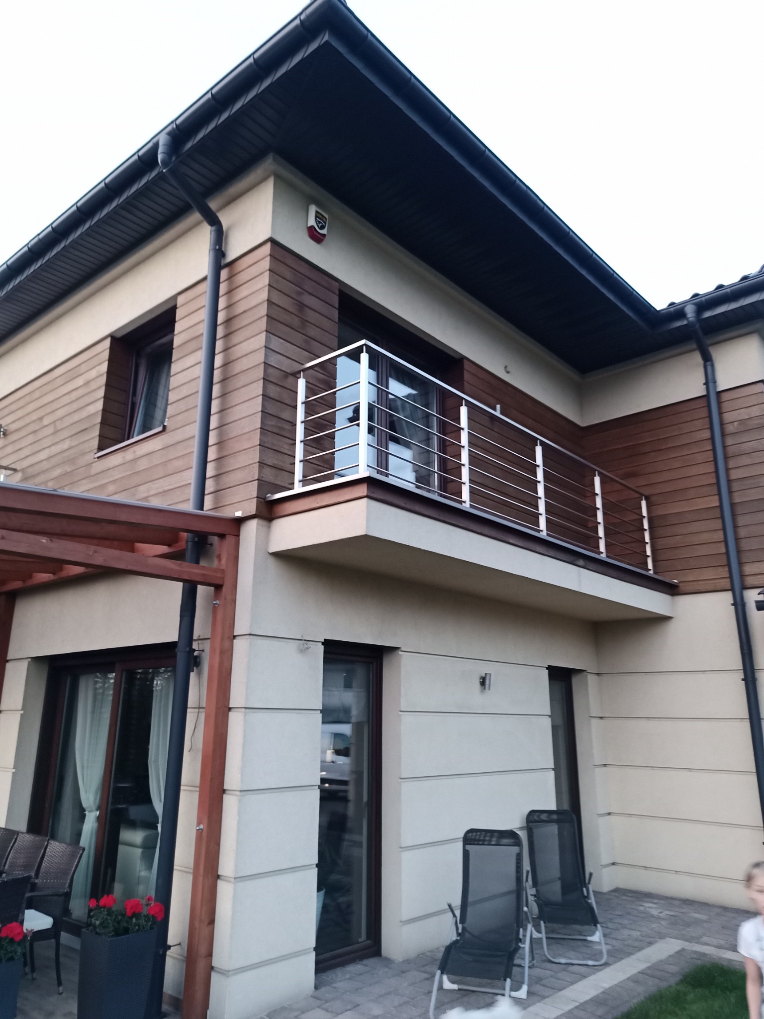 Dwupiętrowy dom z beżową elewacją i drewnianą okładziną, balkon z chromowanymi barierkami, widoczne rynny i elementy wykończeniowe dachu