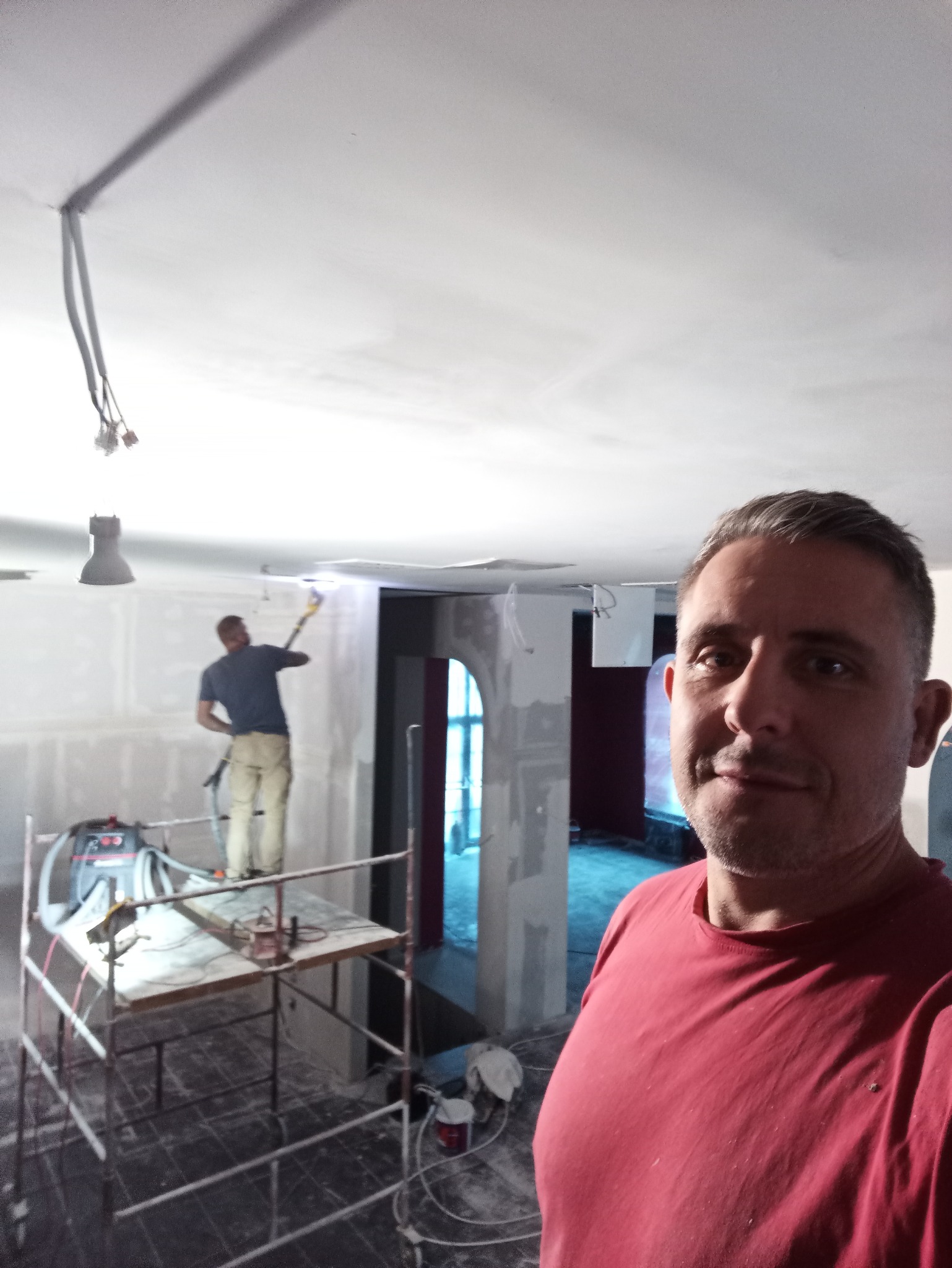 Mężczyzna robiący selfie na tle remontowanego wnętrza: szpachlowanie sufitu, rusztowanie i sprzęt do szlifowania widoczne w tle.