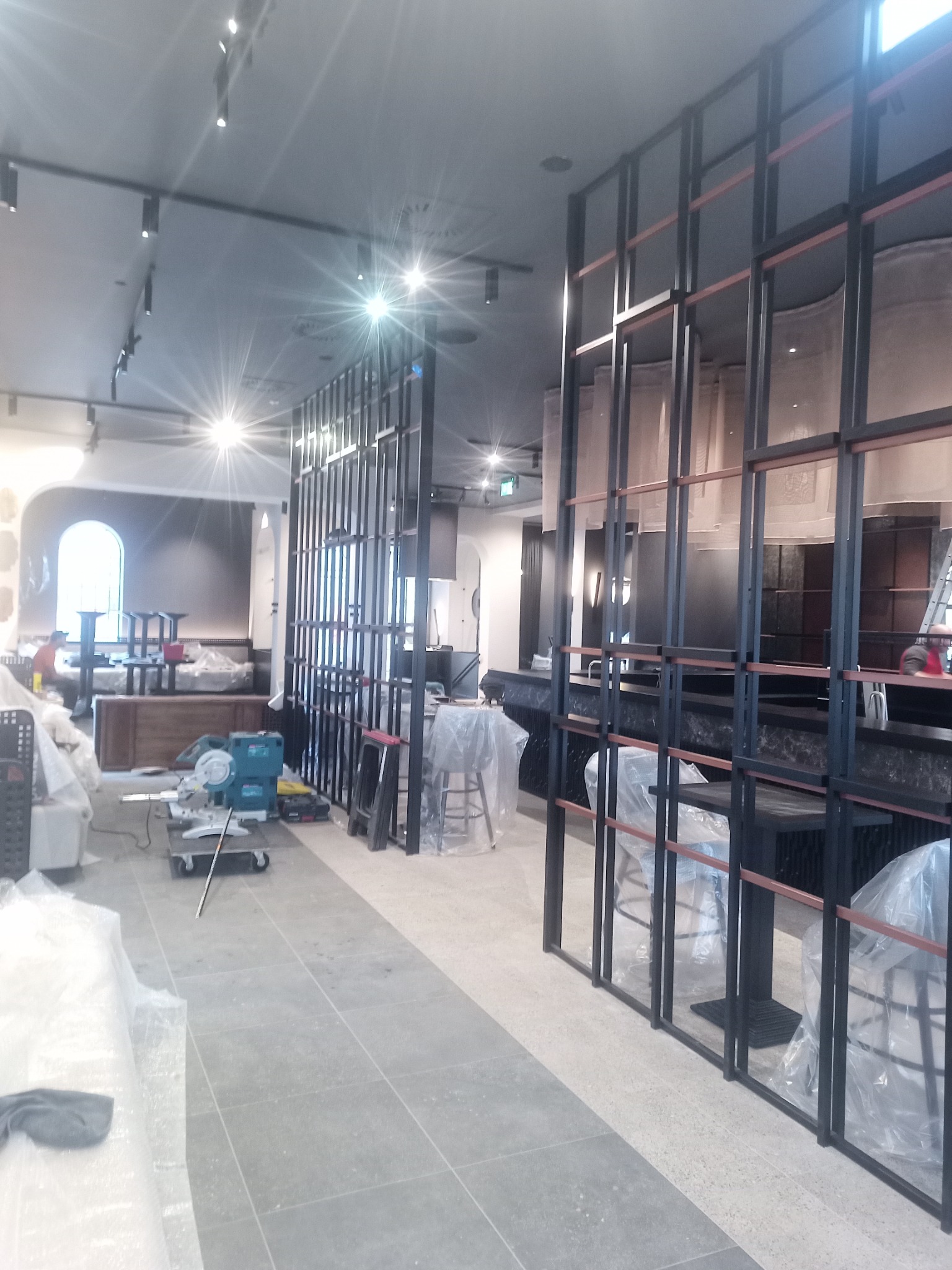Wnętrze lokalu komercyjnego w trakcie wykańczania: widoczne metalowe ramy działowe, krzesła barowe zabezpieczone folią, narzędzia budowlane oraz posadzka z płytek
