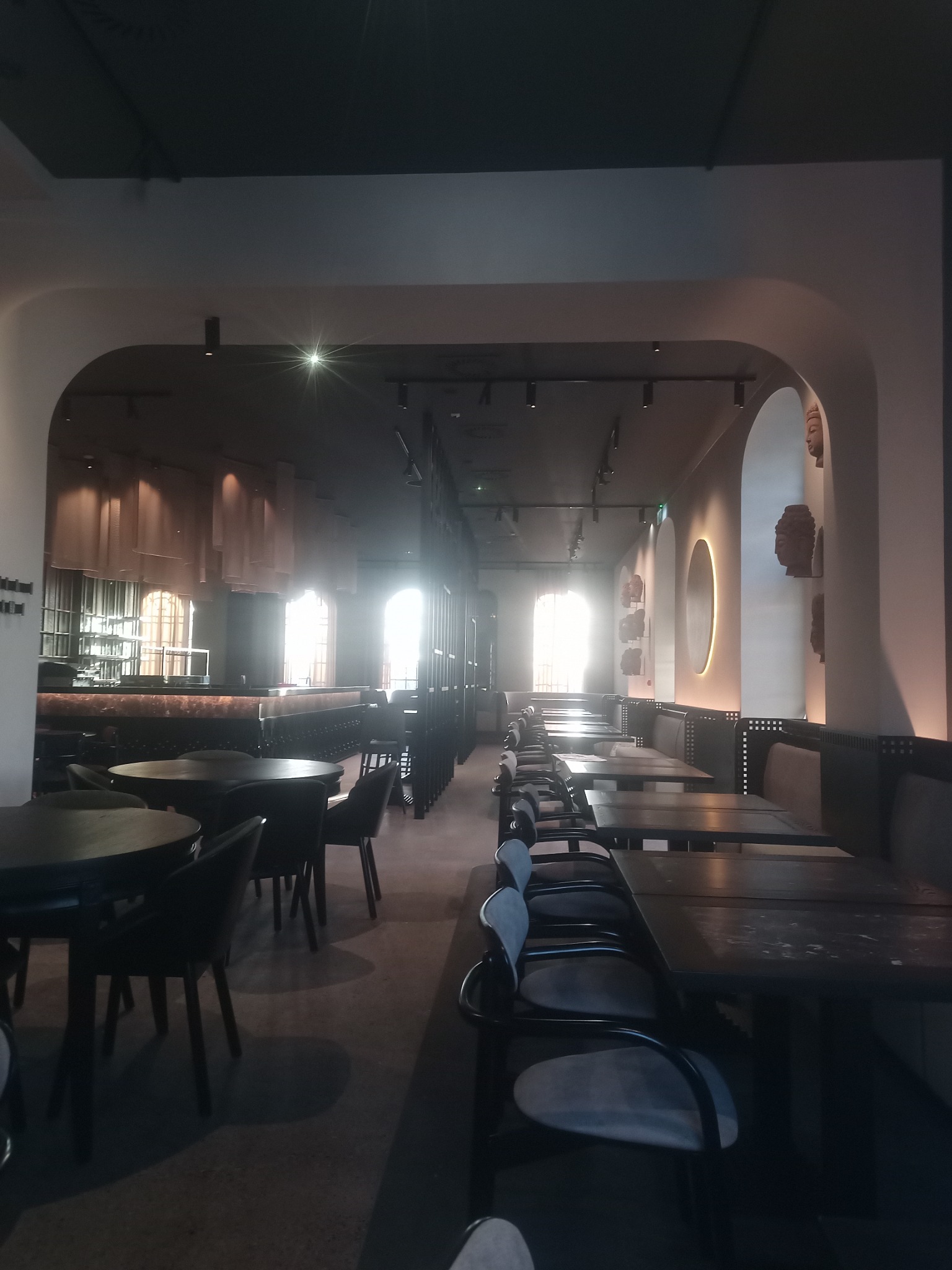 Wnętrze restauracji z rzędem stolików i krzeseł, oświetlone promieniami słonecznymi wpadającymi przez okna, dekorowane rzeźbami głów na ścianach i podwieszanymi elementami dekoracyjnymi.