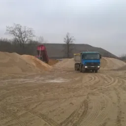 Ciężarówka Mercedes-Benz z niebieską kabiną jedzie po piaszczystym terenie, za nią hałdy piasku i druga ciężarówka z podniesioną czerwoną skrzynią ładunkową, pochmurne niebo.